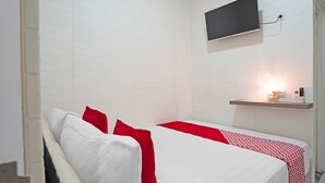 Une chambre d’hôtel avec un lit blanc, des oreillers rouges, un téléviseur fixé au mur et une table de chevet avec une lampe.