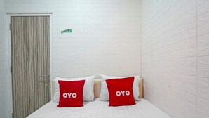 Une chambre d’hôtel avec un lit, deux oreillers rouges portant la marque « OYO », une porte blanche et un mur blanc à texture.