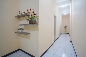 Un couloir aux murs et au sol recouverts de carreaux blancs, agrémenté d’étagères grises fixées au mur, sur lesquelles sont disposées des plantes et des fleurs en pot.