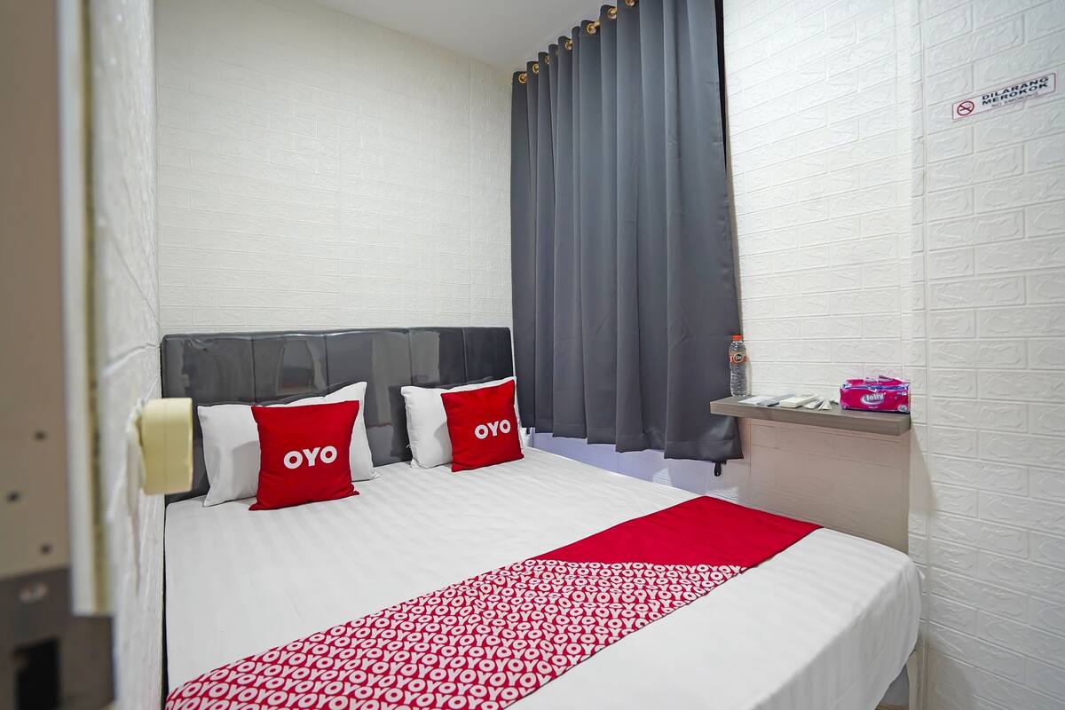 Habitación de hotel con cama, cortinas grises, pared de ladrillo blanco y repisa con artículos de aseo.