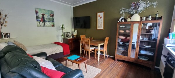 A living room with a sofa, a dining table with chairs, a TV mounted on the wall, and a cabinet with glass doors.