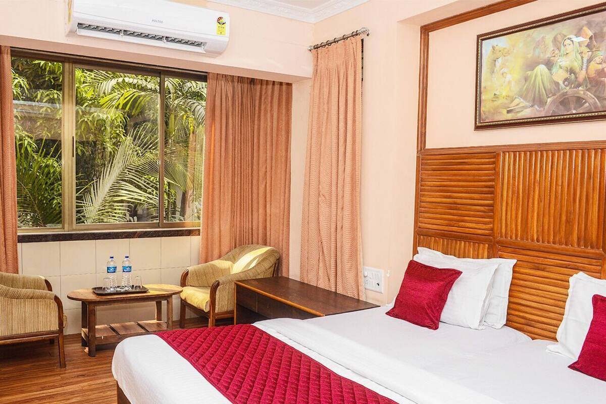 A hotel room with a bed, two chairs, a small table with water bottles, a window with curtains, and a painting on the wall.