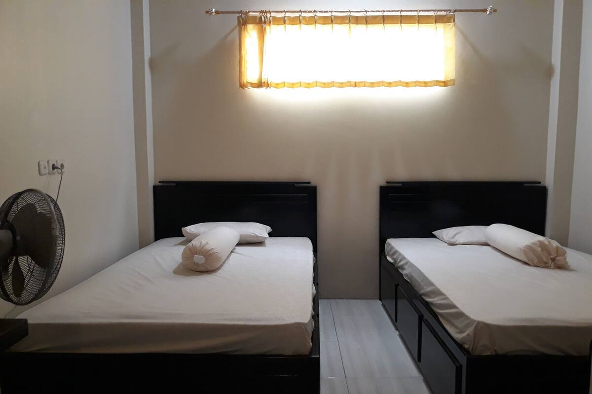 Een kamer met twee bedden, een ventilator en een raam met gordijnen.