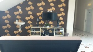 Een kamer met behang met een patroon, een bevestigde televisie, een klein wastafeltje en een kast met diverse spullen.