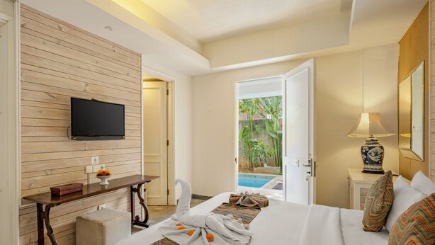 A hotel room with a wooden wall, a flat-screen TV, a desk with a vase, a bed with patterned pillows, and a view of a swimming pool. - The Kanjeng Suites Petitenget (Keroboken Kelod)