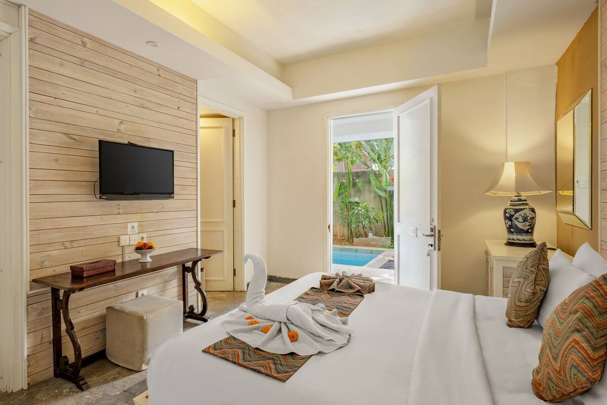 Une chambre d’hôtel avec un mur en bois, un téléviseur à écran plat, un bureau agrémenté d’un vase, un lit avec des coussins à motifs et une vue sur une piscine.
