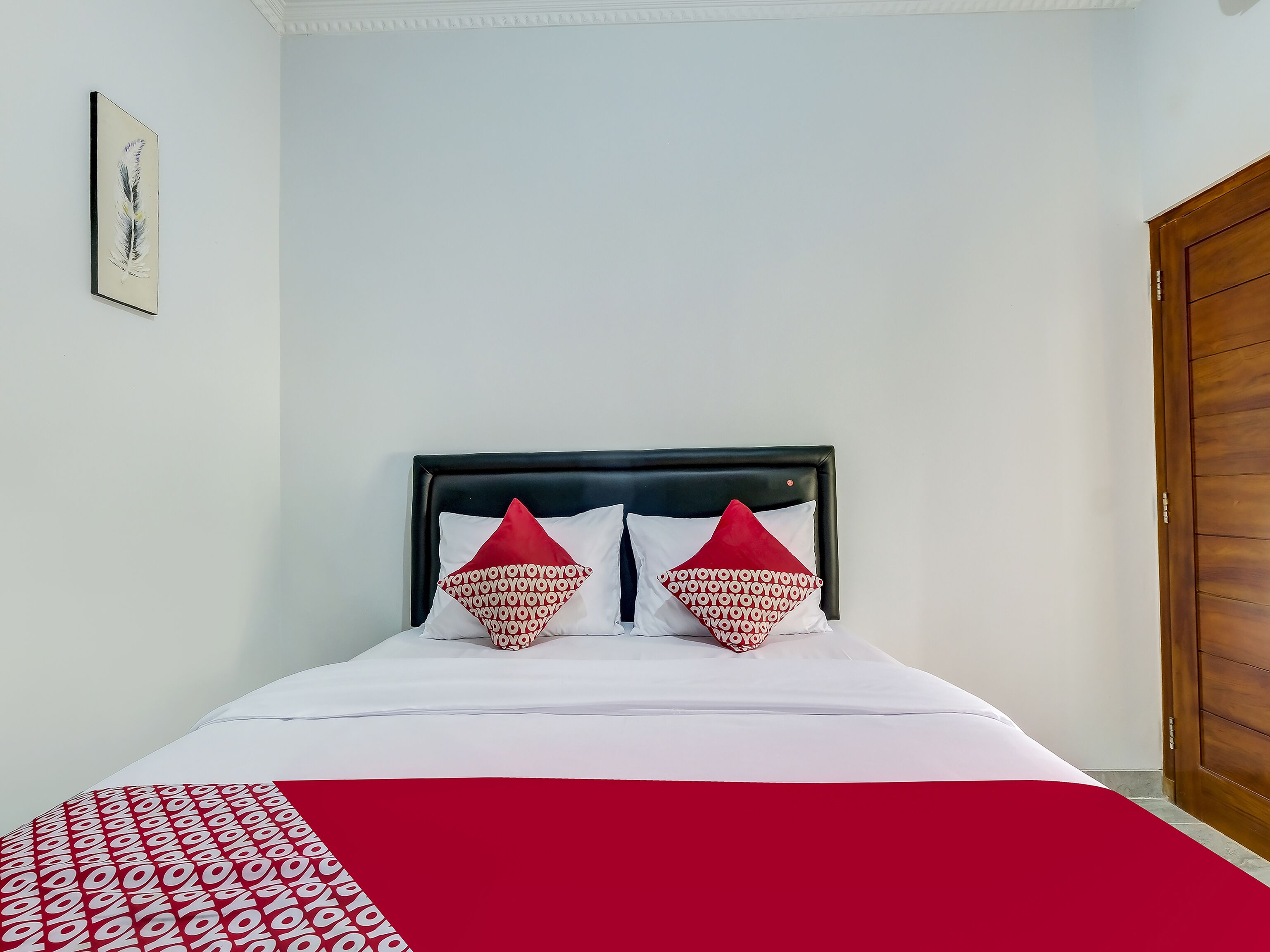 Un lit recouvert d’une couvre-lit à motifs rouges et blancs, deux oreillers rouges ornés du même motif, et un tableau encadré accroché au mur.