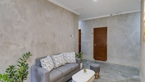 A modern living room with a grey sofa, a white coffee table, and two grey ottomans. There are potted plants and a wooden door in the background. - Capital O 90983 Harmony Easy Stay (Depok)