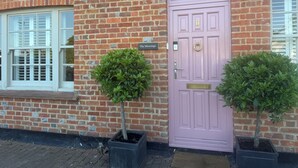A brick building with a pink door, two potted trees, and a window with shutters.