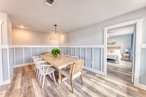 A dining area with a wooden table and white chairs, a hanging light fixture, and a plant. - Beachfront |Cargo Elevator | Steps to Beach | Baby+Beach Gear | Lawn+Board Games (Long Beach)