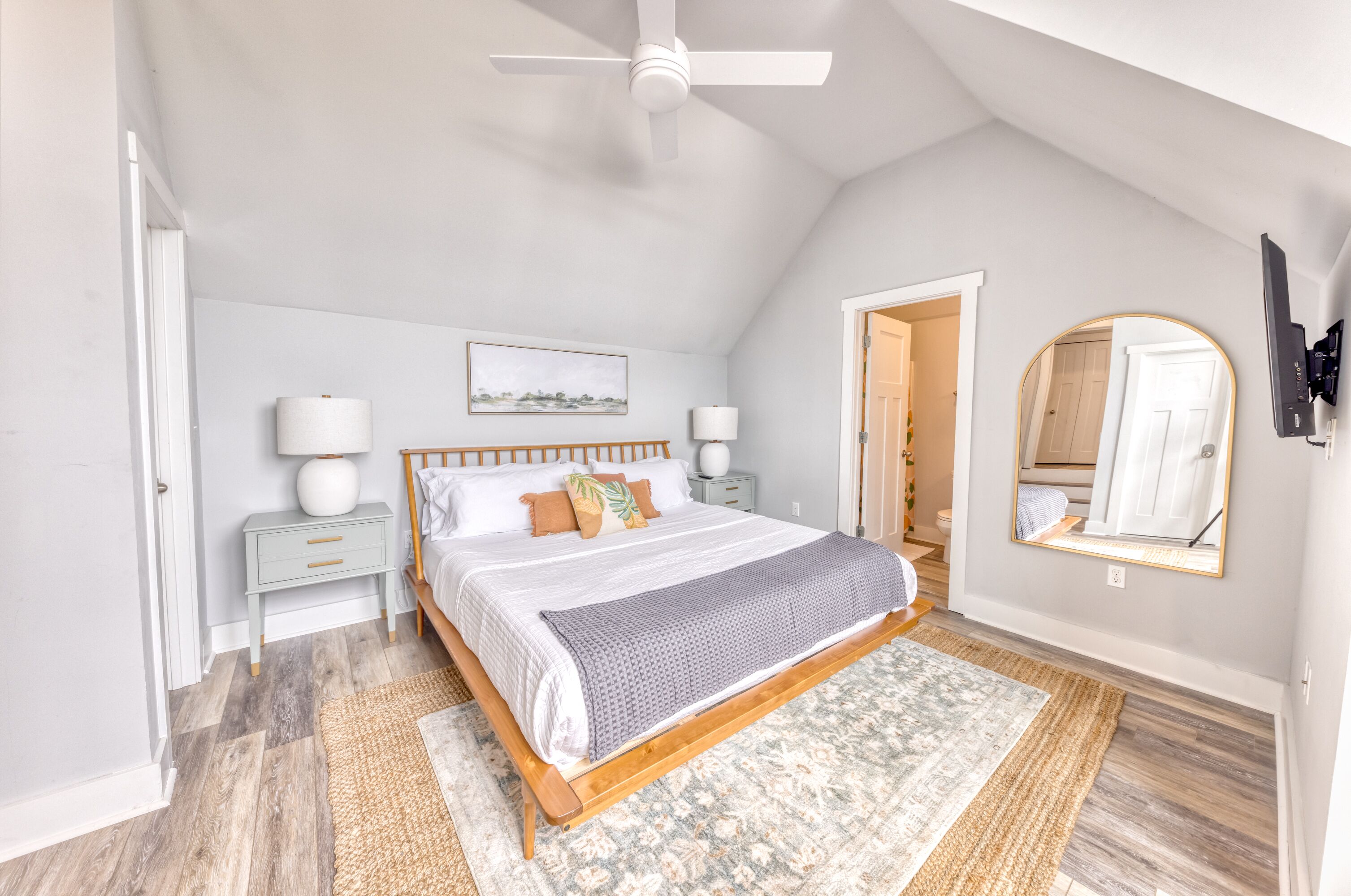 A bedroom with a bed, nightstands, a ceiling fan, a large mirror, and a door leading to another room.