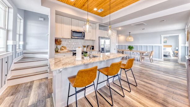 A modern kitchen with a wooden ceiling, white cabinets, a central island with bar stools, and a view into the dining area. - Beachfront |Cargo Elevator | Steps to Beach | Baby+Beach Gear | Lawn+Board Games (Long Beach)