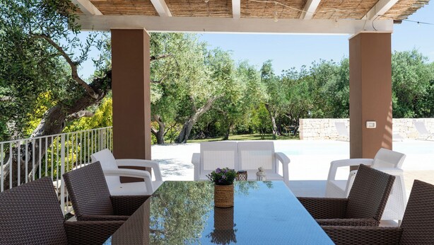 Un patio exterior con mobiliario de mimbre, una mesa de vidrio y vistas a una piscina y árboles.