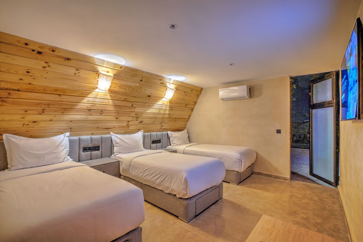 Habitación de hotel con tres camas, paneles de madera en las paredes, un equipo de aire acondicionado y una puerta corrediza de vidrio que da a un espacio exterior.