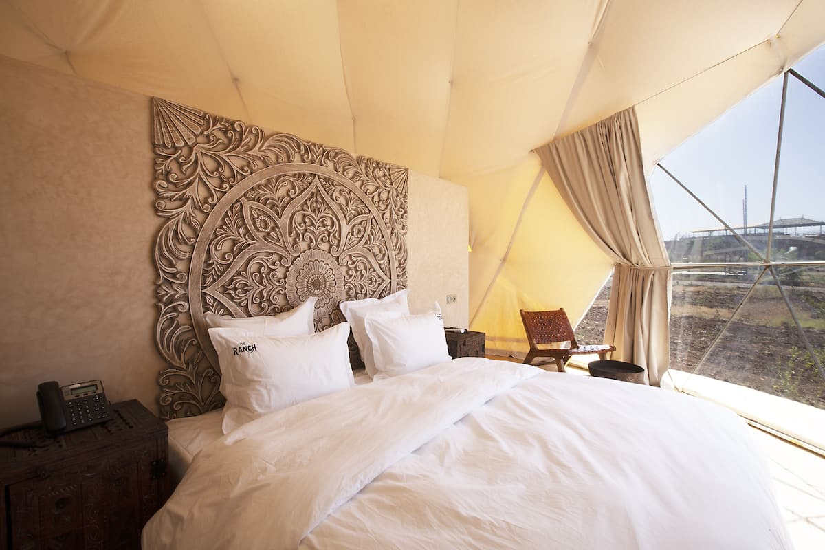 Ein Schlafzimmer mit einem großen Bett, einem hölzernen Kopfteil, einem Nachttisch mit Telefon und Blick ins Freie durch ein Fenster.