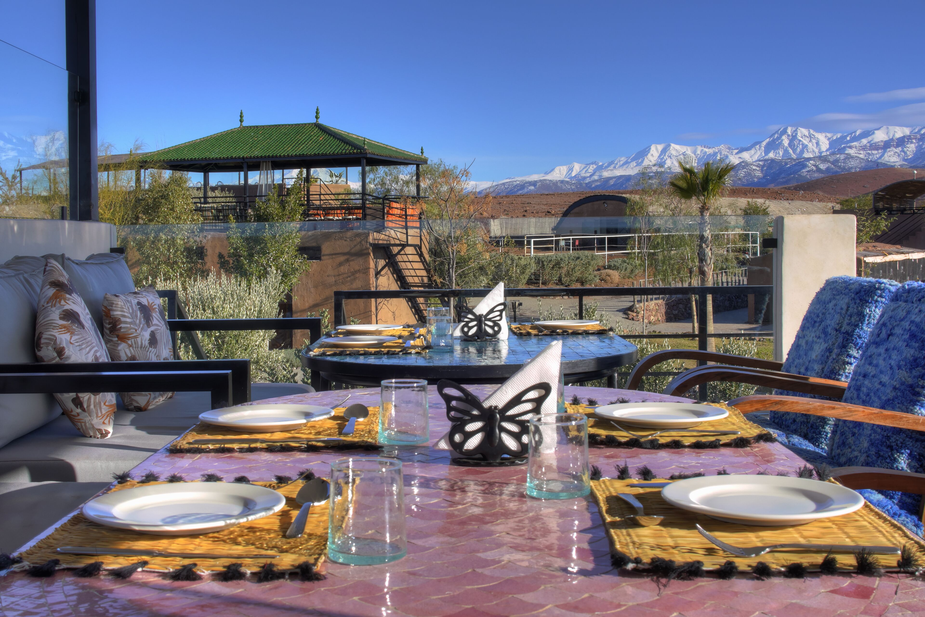 Un área de comedor al aire libre con vistas a montañas nevadas, una zona de estar techada y una mesa dispuesta con platos y vasos.