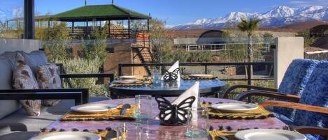 Un área de comedor al aire libre con vistas a montañas nevadas, una zona de estar techada y una mesa dispuesta con platos y vasos.