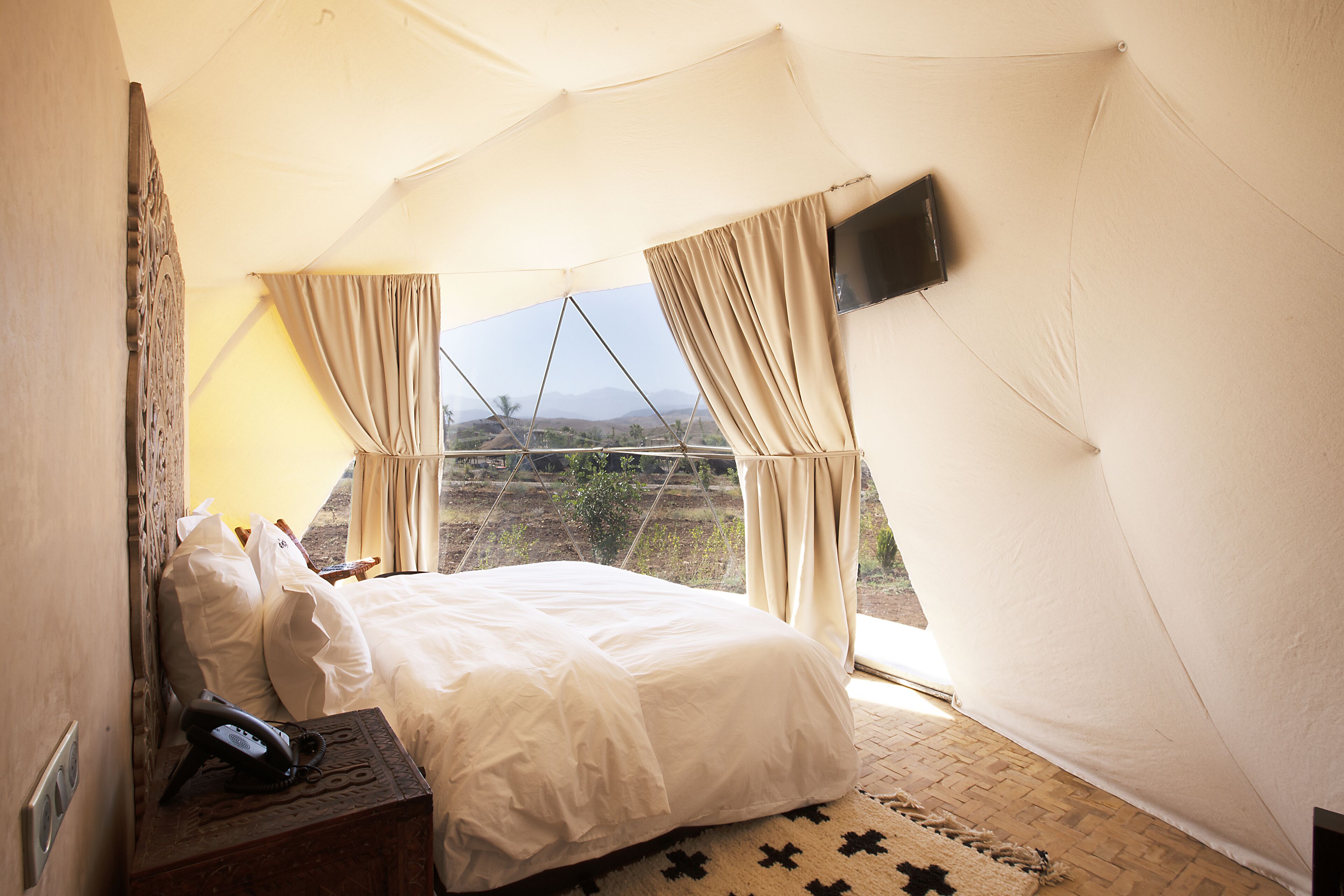 Habitación con tienda de campaña, cama, cortinas, televisor montado y teléfono sobre una mesita de noche de madera.