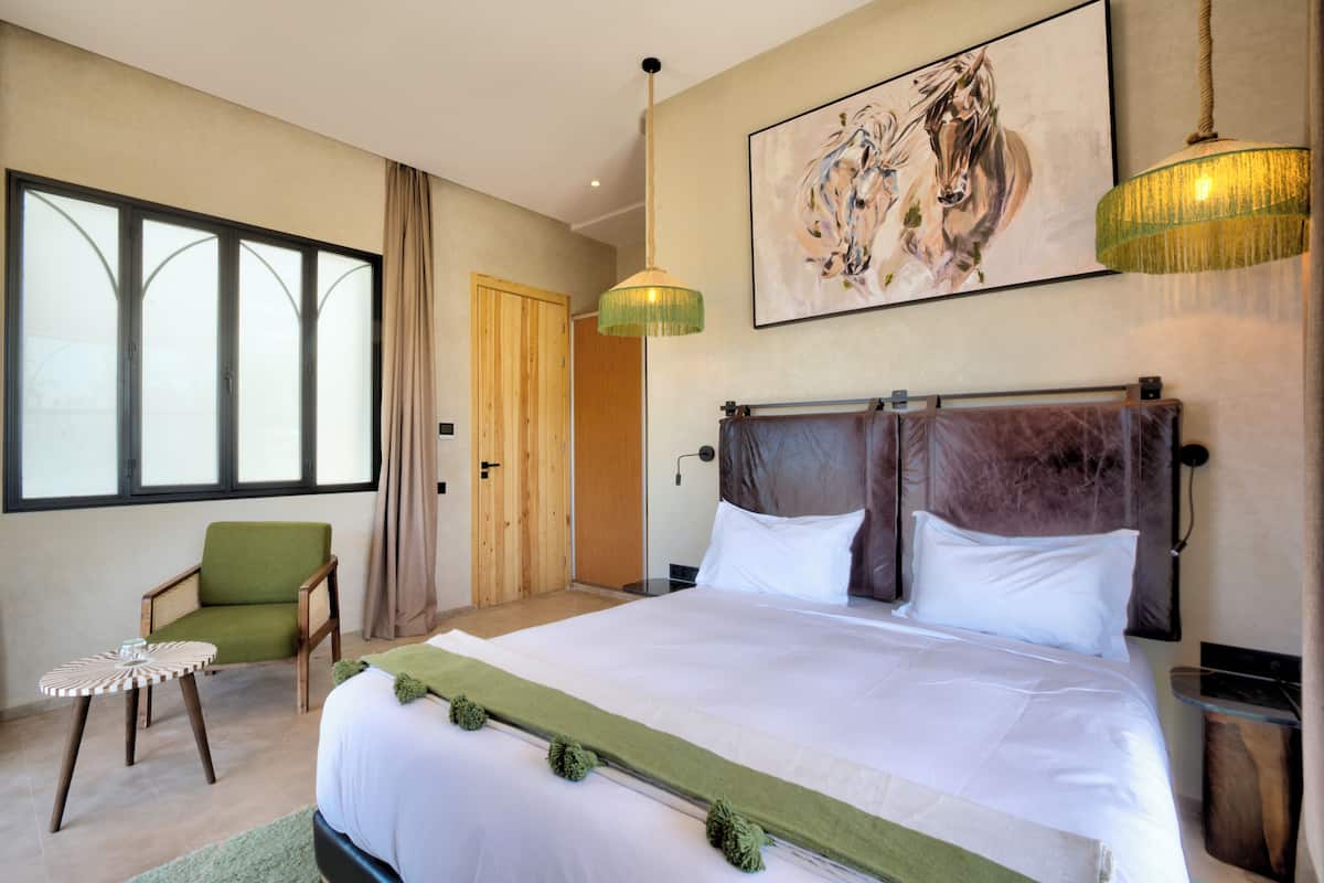 Ein geräumiges Schlafzimmer mit einem großen Bett, einem grünen Sessel, einem runden Beistelltisch und einem großen Fenster mit Vorhängen.