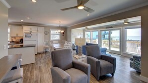 A spacious living area with a dining table, comfortable seating, a ceiling fan, and large windows offering a view of the ocean.