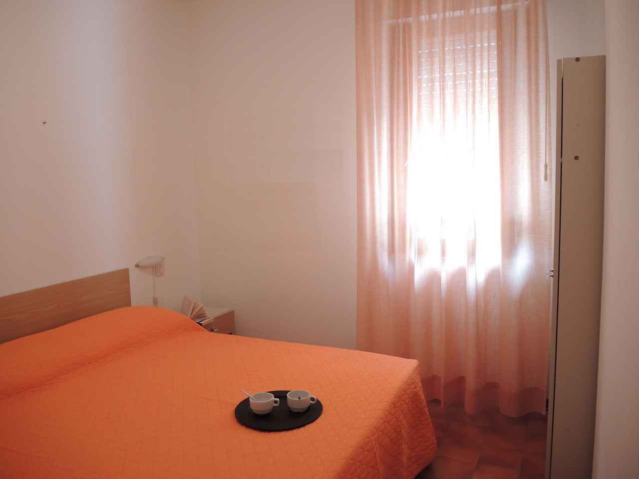 A hotel room with a bed, a small table with two cups, a door, and a window with curtains.