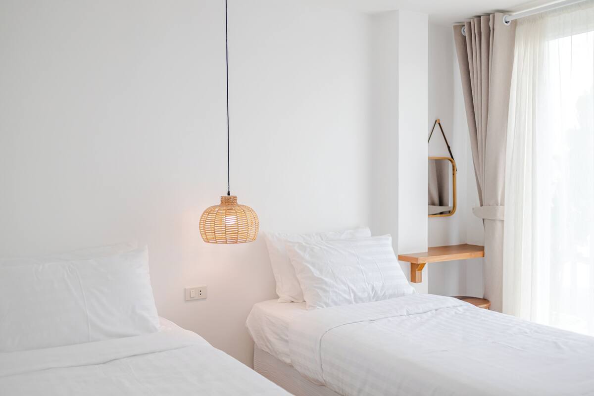 A bedroom with two beds, a hanging light fixture, a wooden shelf, and a window with curtains.