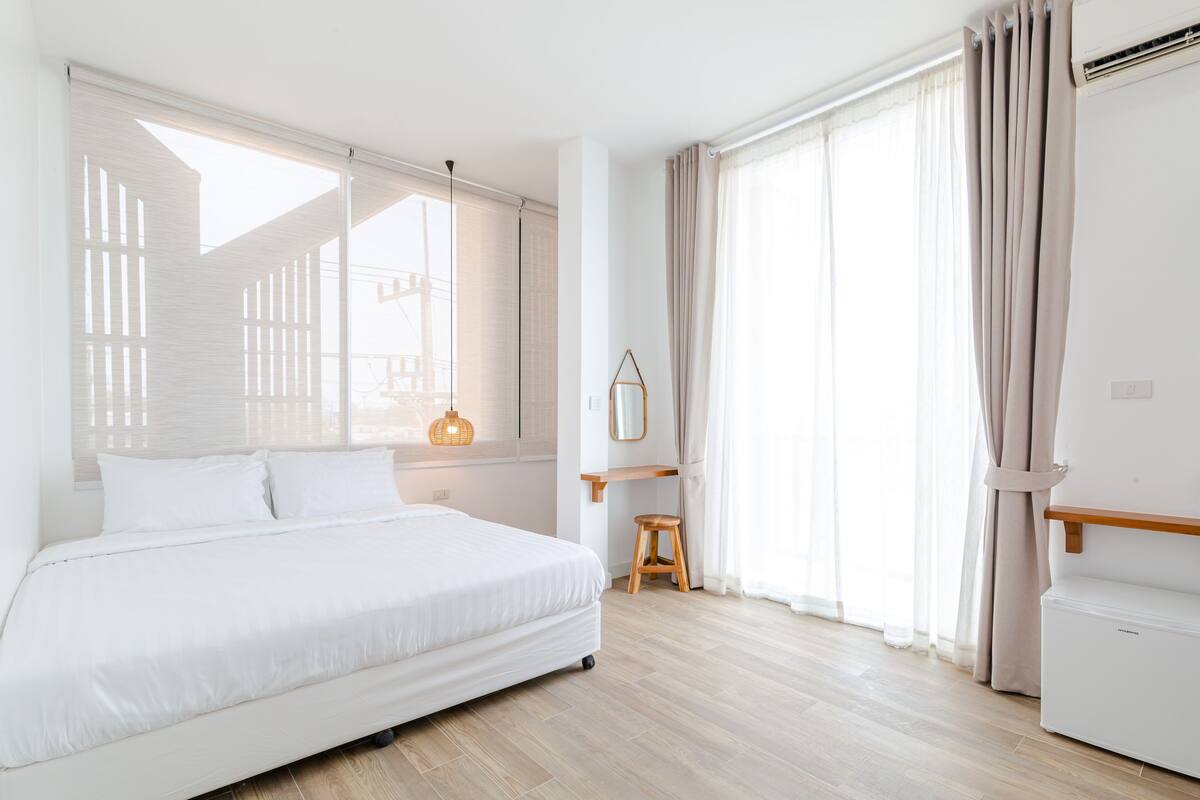 A modern hotel room with a large bed, a wooden floor, a small stool, and a window with curtains.