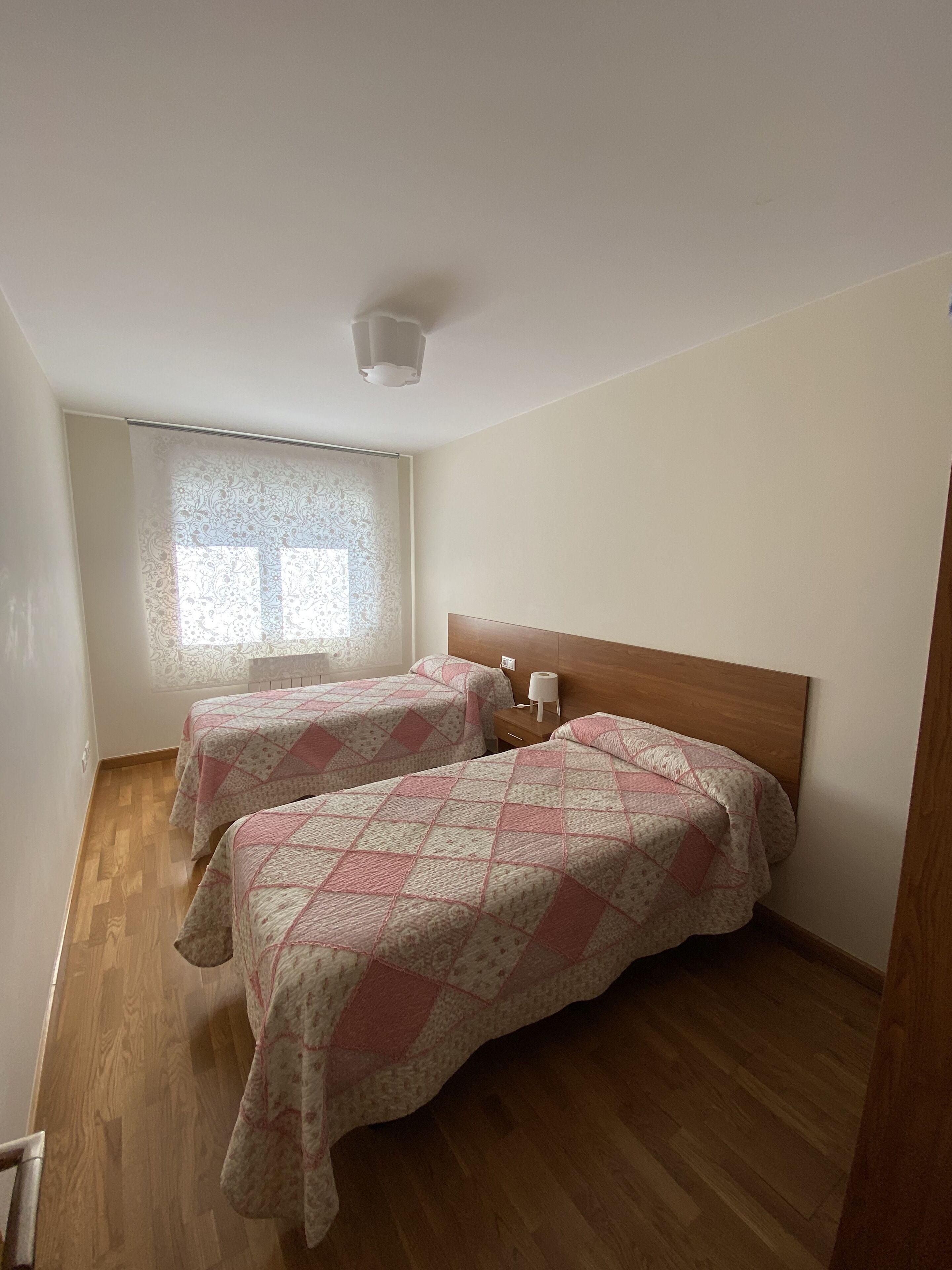 A room with two beds, a wooden headboard, a window with patterned curtains, and a ceiling light.