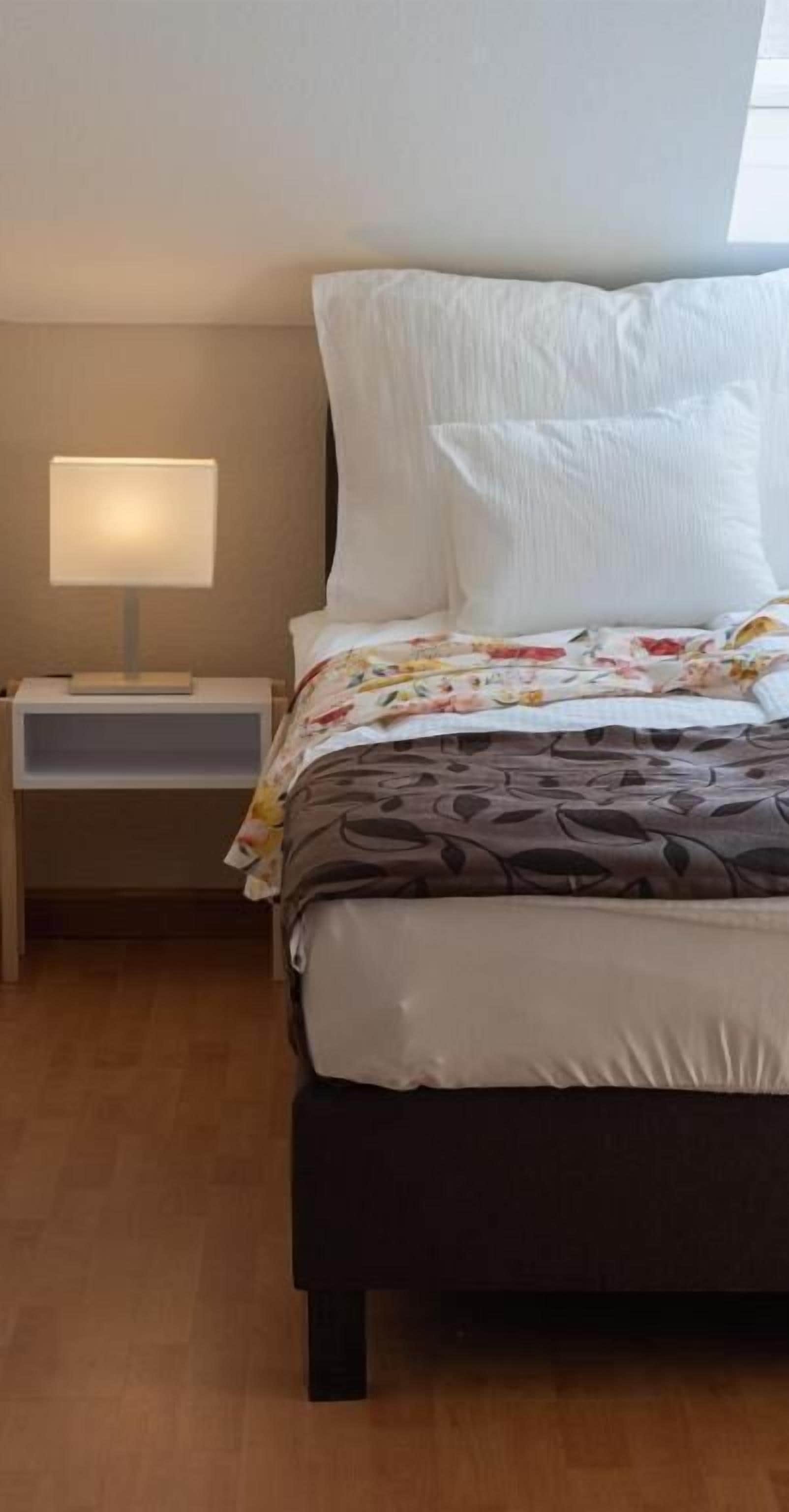 A neatly made bed with a floral duvet, a bedside lamp, and a wooden floor.