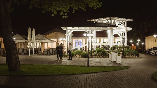 A well-lit outdoor area with a covered seating area, a paved walkway, and a building with a distinctive architectural design. - Budapest Airport Eprespark Hotel (Vecsés)
