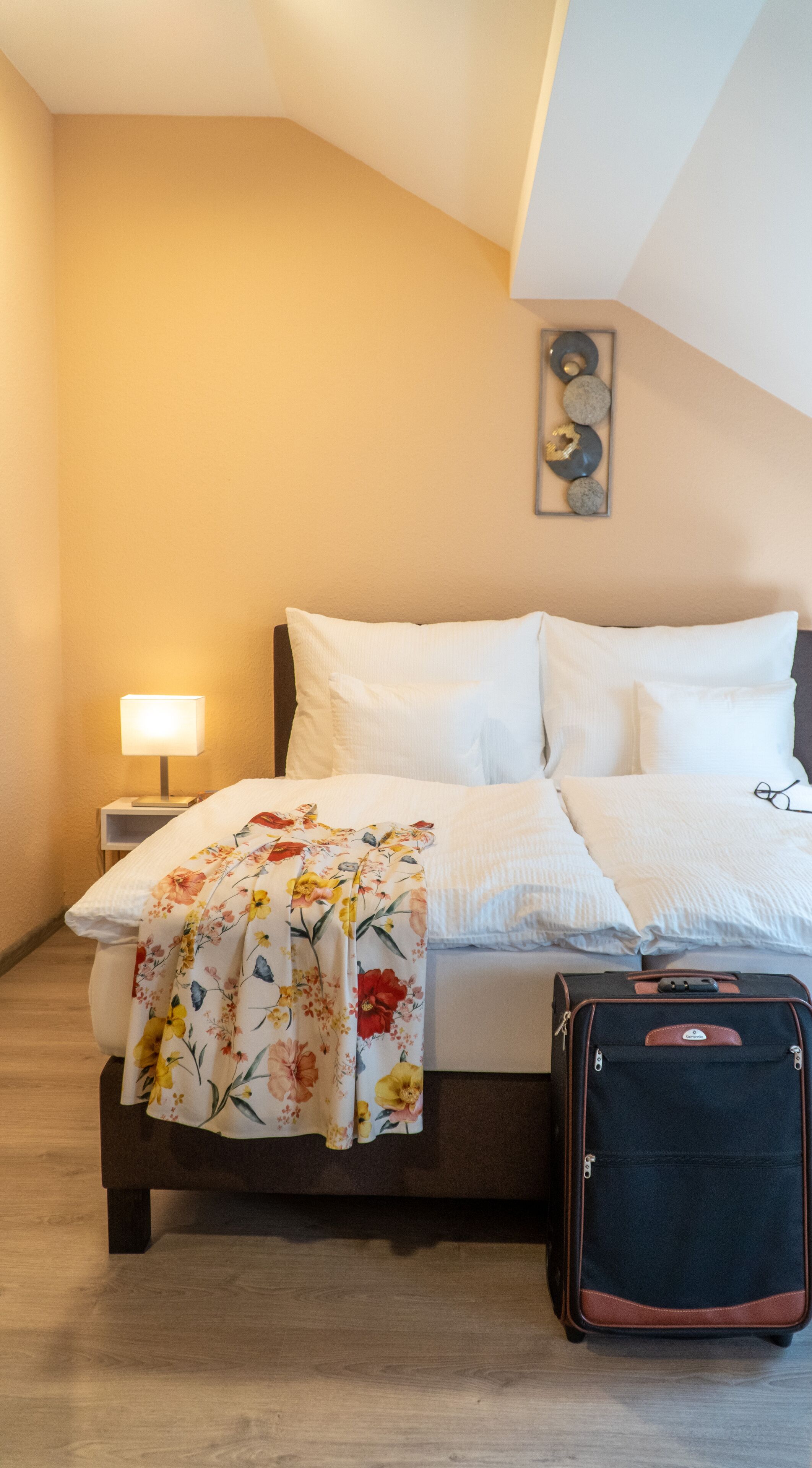 A neatly made bed with a floral blanket, a bedside lamp, and a suitcase on the floor.