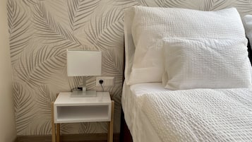 Una cama bien hecha con ropa blanca, una mesita de noche con lámpara y un enchufe.
