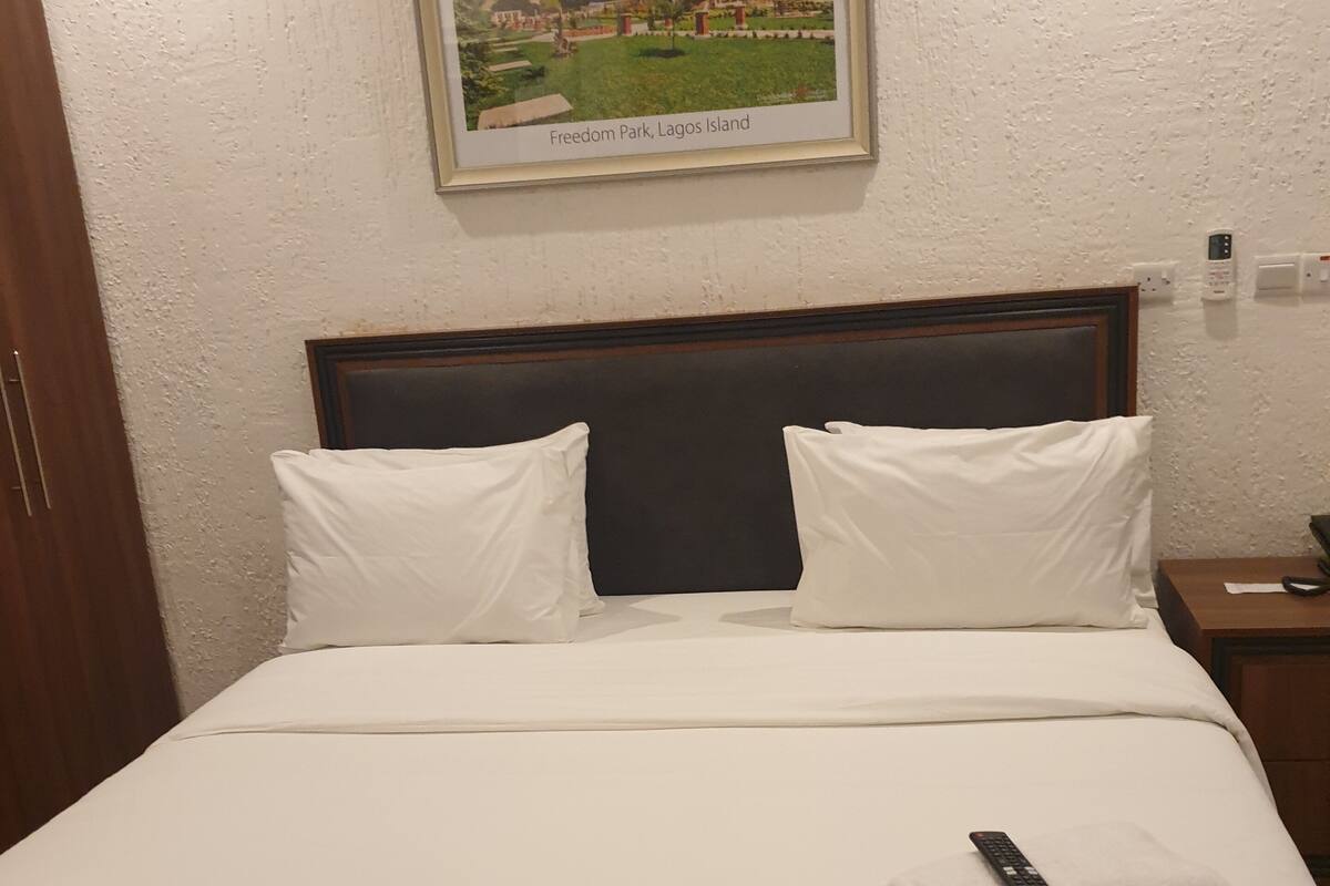 Ein Hotelzimmer mit Bett, Kissen, einer Fernbedienung und einem gerahmten Bild vom Freedom Park auf Lagos Island.