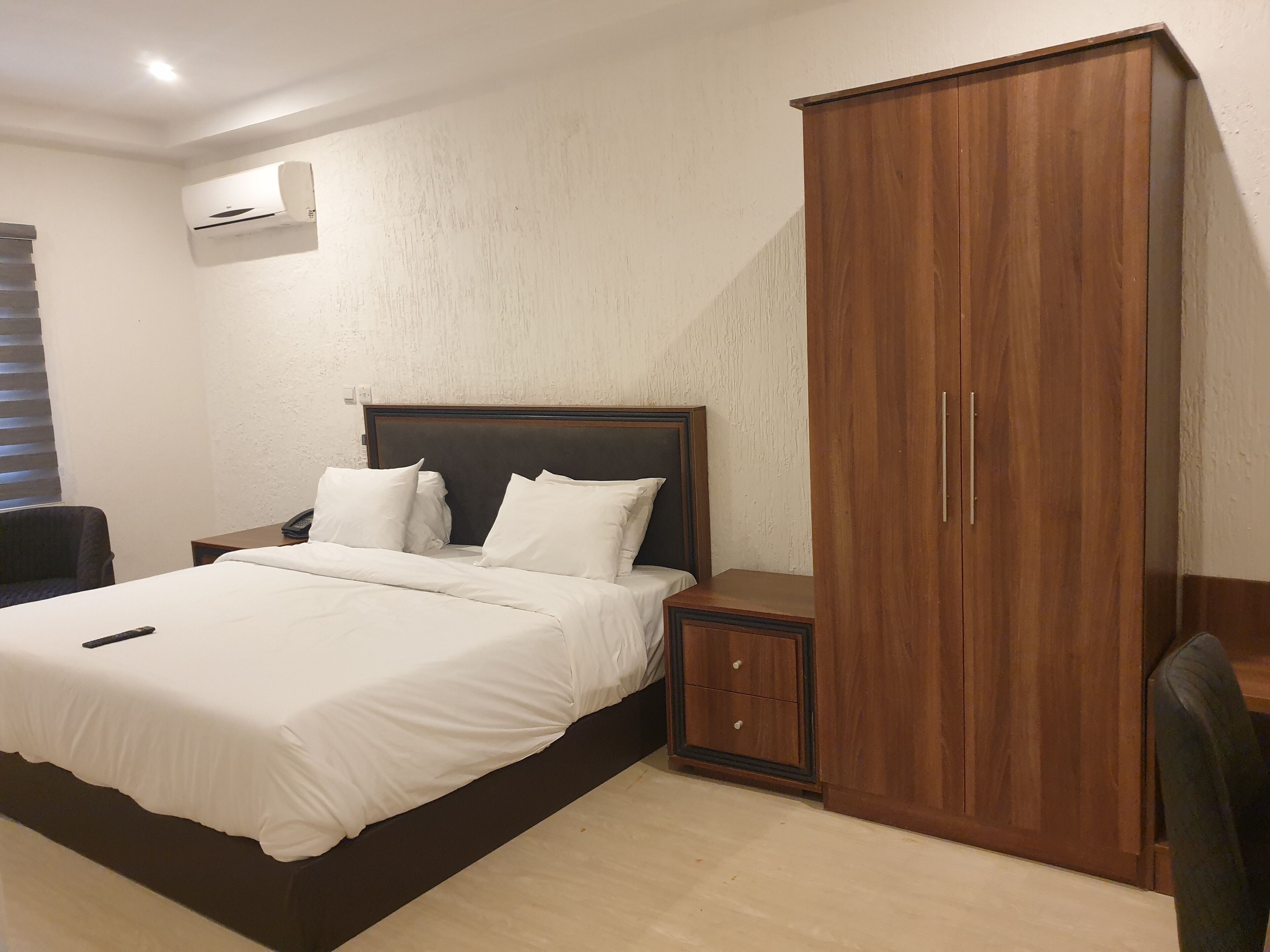 Una habitación de hotel con una cama grande, un armario de madera, una mesita de noche con lámpara y un equipo de aire acondicionado en la pared.