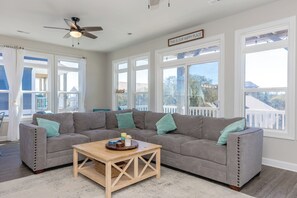 A spacious living room with a grey sectional sofa, a wooden coffee table, and large windows offering a view of the outside.