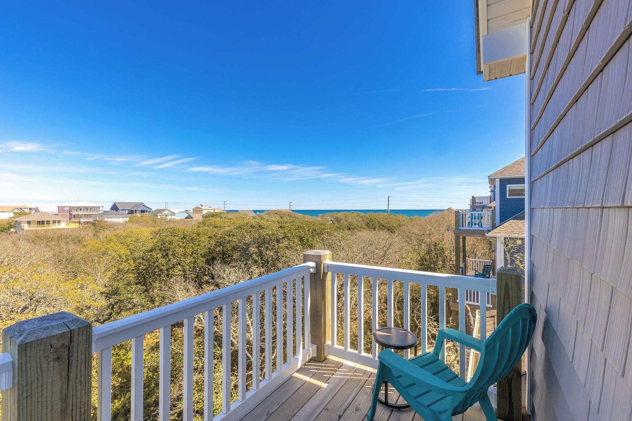 A wooden deck with a white railing, a small table, and a turquoise chair, overlooking a coastal landscape with houses and a clear sky.