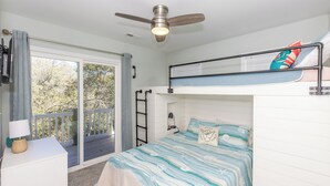 Quarto com cama beliche, ventilador de teto, escrivaninha com luminária e vista para o exterior por uma porta de correr.