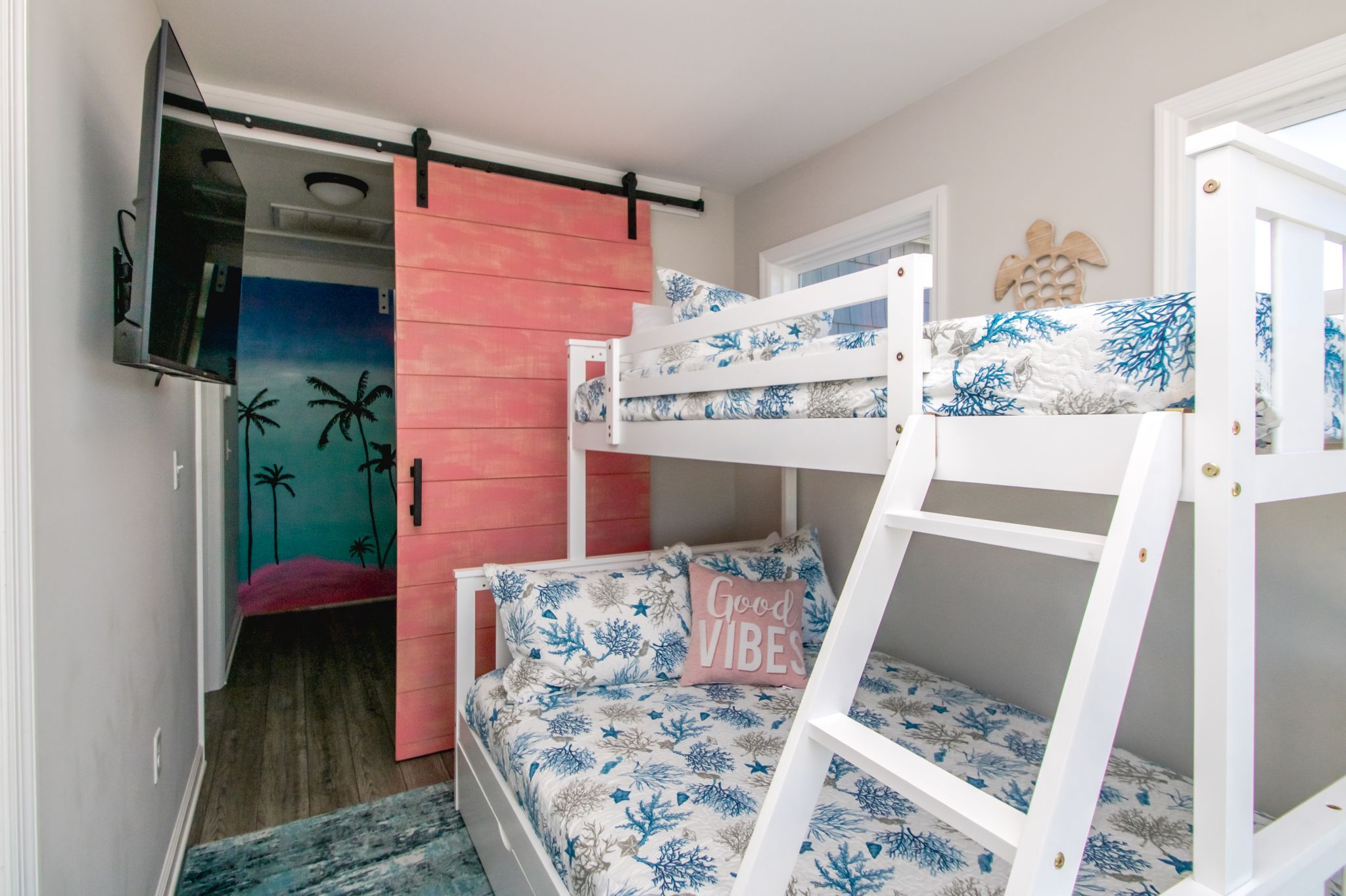 A bunk bed room with a television mounted on the wall, a pink sliding door, and a bed with a 'Good Vibes' pillow.