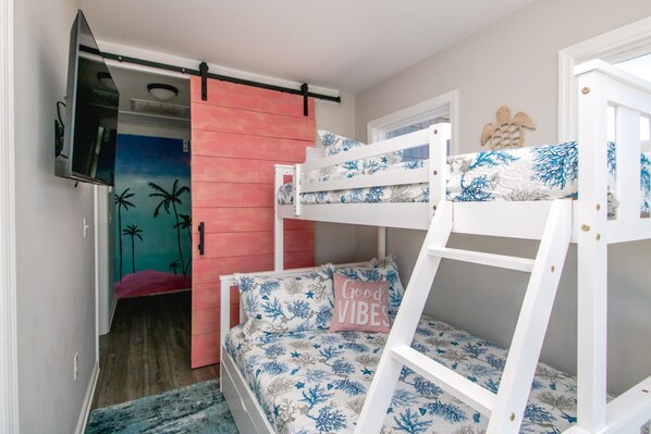 Habitación con literas, televisor montado en la pared, una puerta corredera rosa y una cama con almohada que dice 'Good Vibes'.