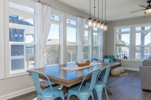 A dining area with a wooden table and blue chairs, large windows, and a ceiling fan.