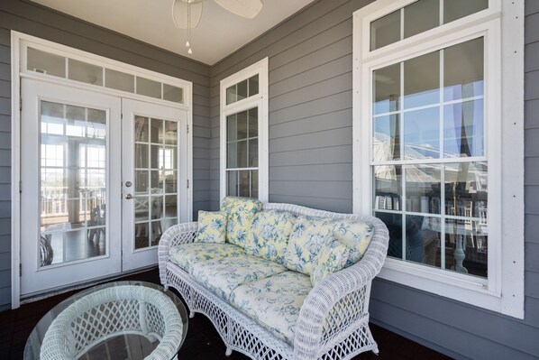 A covered porch with a white wicker sofa and chairs, large windows, and a ceiling fan.