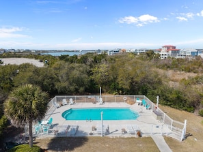 A swimming pool with a fenced deck, surrounded by greenery and overlooking a coastal area with houses.