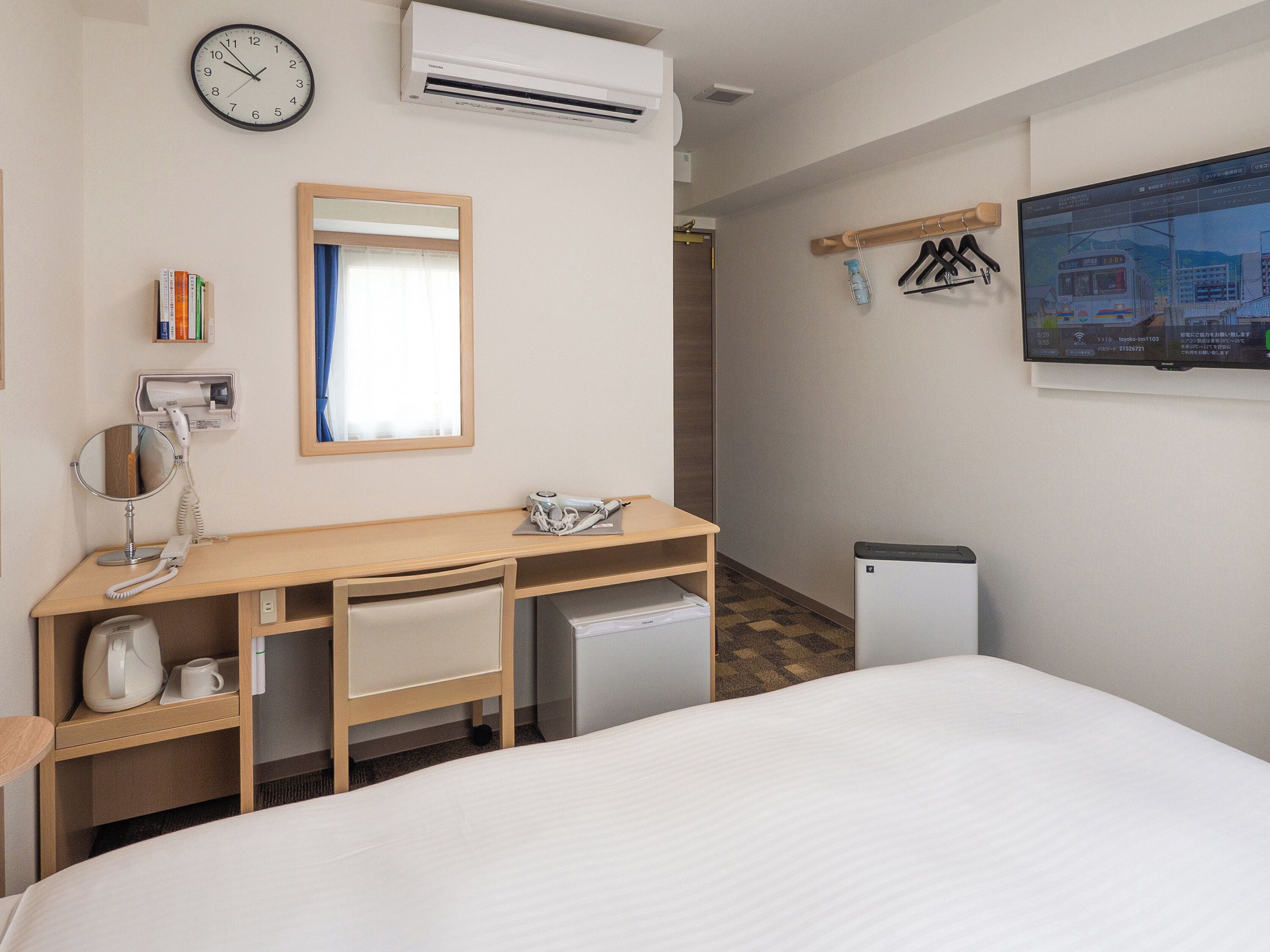A hotel room with a bed, a desk with a mirror, a small kitchenette, a wall-mounted TV, and a clock.