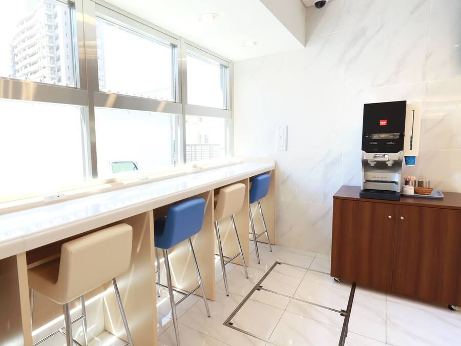A modern bar area with a long counter, three bar stools, a coffee machine, and a view of the city through large windows.