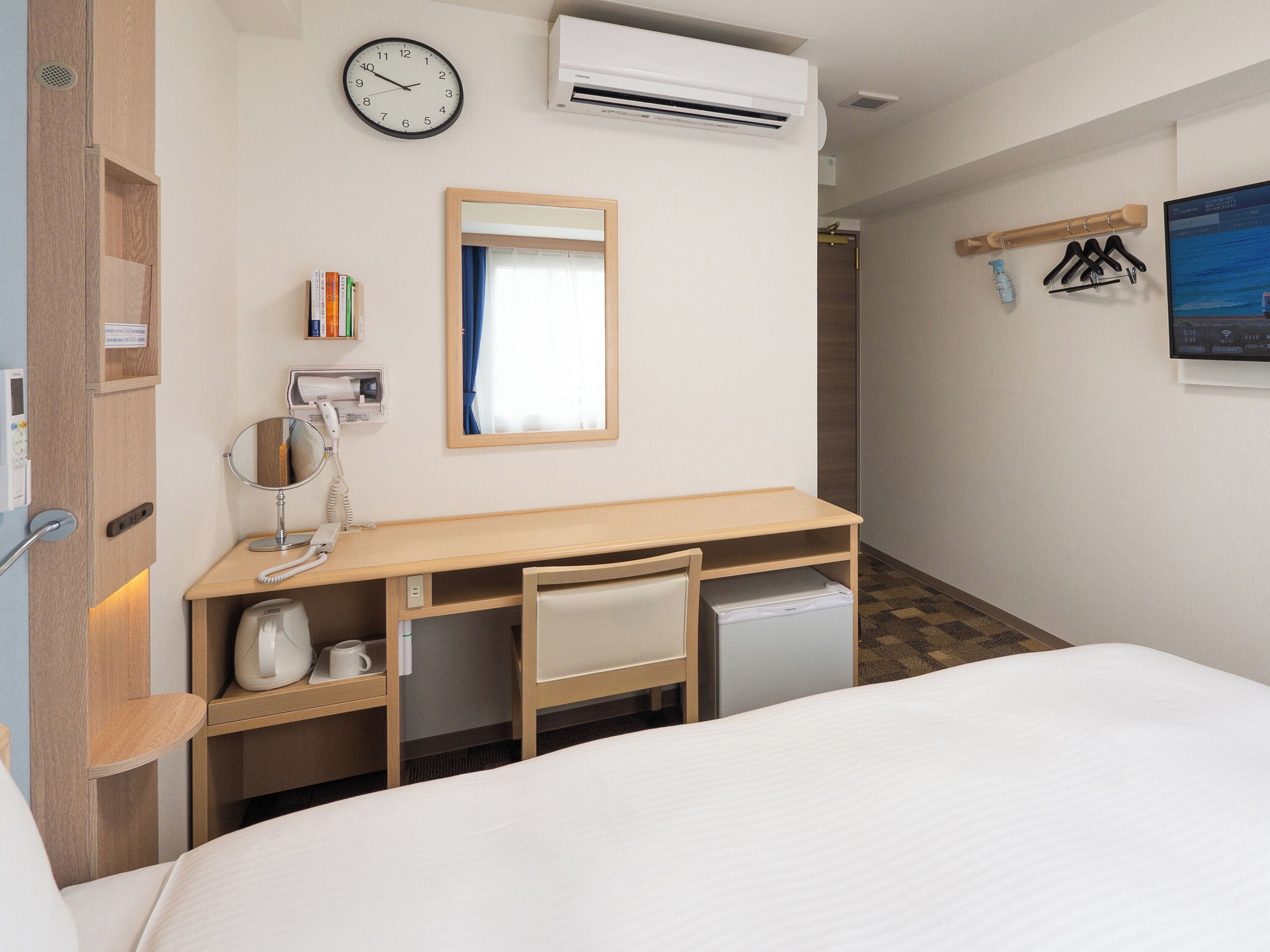Habitación de hotel con cama, escritorio con silla, espejo, reloj, aire acondicionado, refrigerador pequeño y ventana con cortinas.