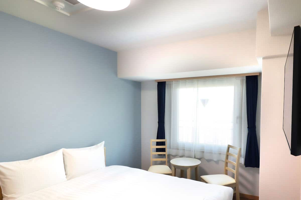 Ein Hotelzimmer mit einem großen Bett, einem kleinen runden Tisch, zwei Stühlen, einem Fenster mit Vorhängen und einem flachbildfernseher an der Wand.