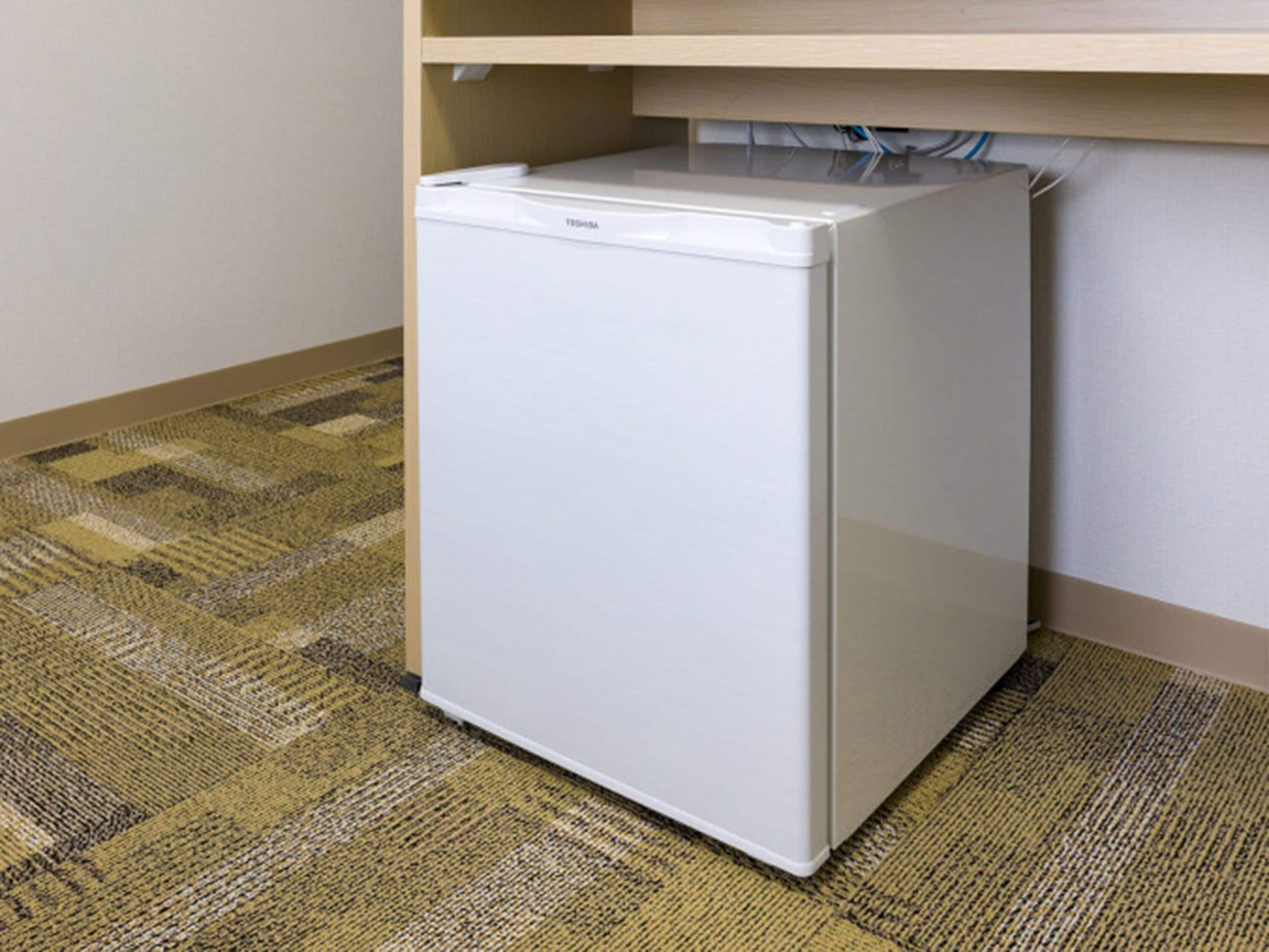 A compact white refrigerator in a corner of a room with a checkered carpet.