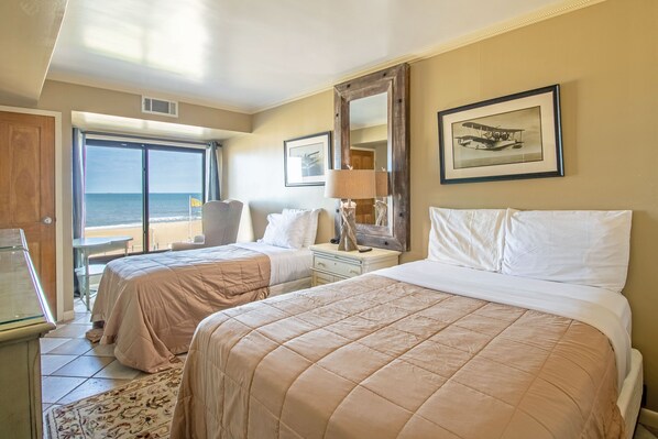 A hotel room with two beds, a desk, a chair, a lamp, and a large window with a view of the sea. - Oceanfront views, Full Kitchen, Prime Location! (Virginia Beach)