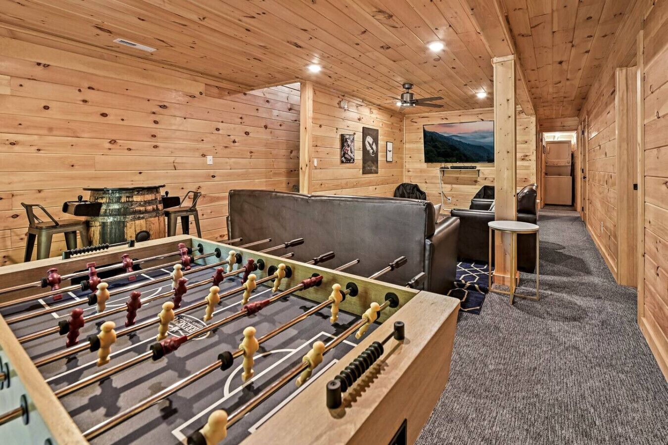 Une pièce au plancher de bois, équipée d’un baby-foot, d’un coin bar et d’un écran de télévision diffusant un paysage pittoresque.
