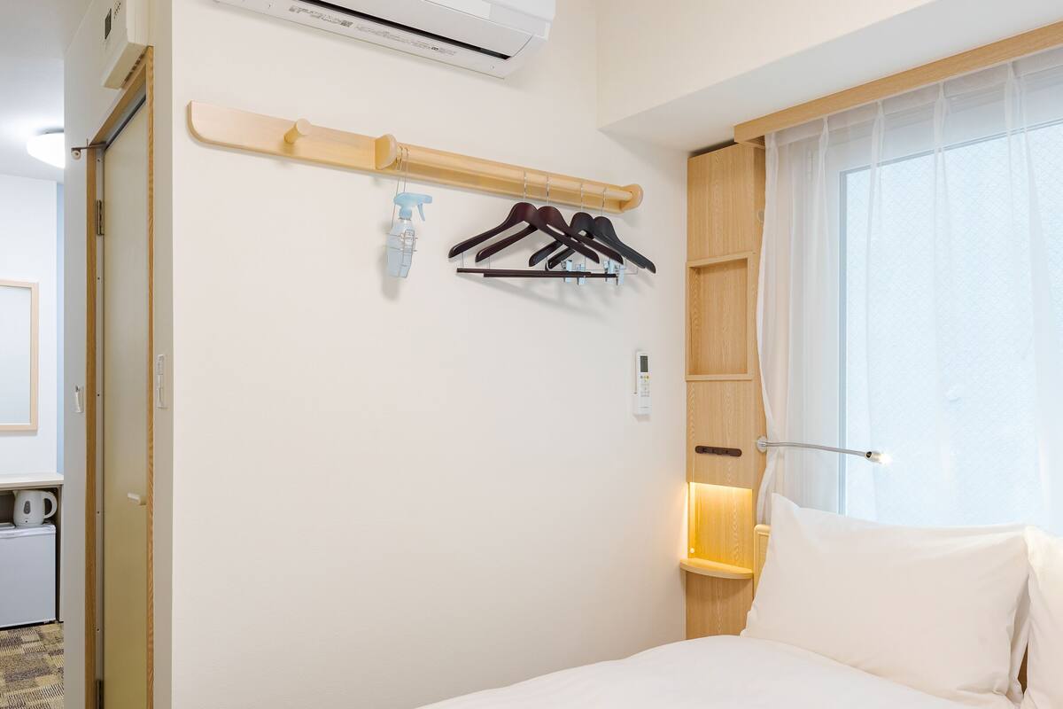 A hotel room with a bed, a wall-mounted air conditioner, a shelf with a spray bottle, and a window with curtains.